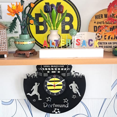 Rezensent zeigt die schwarze Dortmund-Wanduhr aus Vinyl, die an der Wand unter einem Regal hängt.