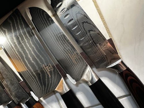 Rezensent zeigt das OKAMI Rei – Santoku Messer mit seiner Damastklinge neben anderen Messern auf einem Messerhalter.