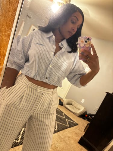 Reviewer in a mirror wearing the pinstriped Overhead Cropped Shirt, pulling it down to show the fit.