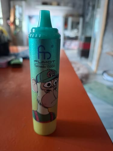 Kunde zeigt einen grün-gelben ORIGINAL RANDM DIGITAL BOX 12000 Vape mit einem Cartoon-Affen-Aufdruck, der auf einem Tisch steht.