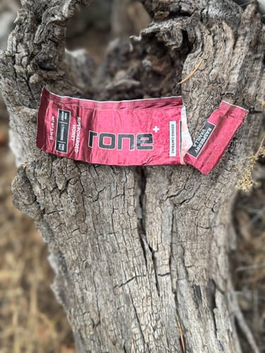 Customer's used Raspberry Vanilla SuperCharged Honey wrapper resting on a gnarled log outdoors.