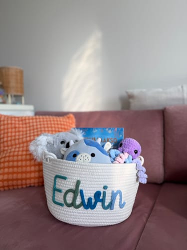 Customer's white personalized knitted basket with a multicolor name 'Edwin', shown on a couch holding several toys.