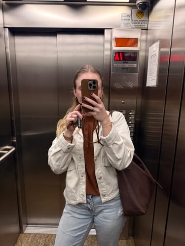 Cliente tirando uma selfie no espelho de um elevador, mostrando a case Nora | Mocha Mousse e sua alça.