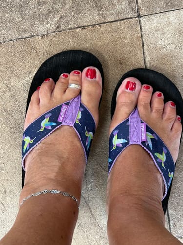 A reviewer's feet shown wearing the Hummingbirds Flip Flops, highlighting the colorful hummingbird pattern on the navy and purple straps.