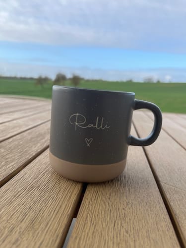 Ein Kunde zeigt eine personalisierte, zweifarbige Tasse mit dem Namen „Ralli“ auf einem Holztisch im Freien.