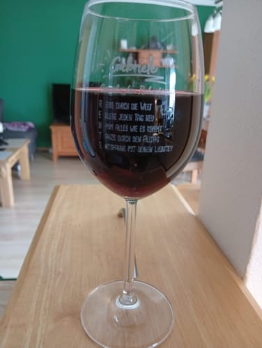 Kundenbild eines personalisierten Weinglases mit Rotwein und einer Gravur, das auf einem Holztisch steht.