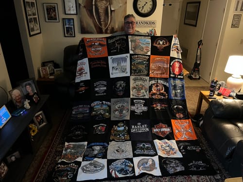 Customer holding up a large Project Repat T-Shirt Quilt made from many different graphic t-shirts in a living room.