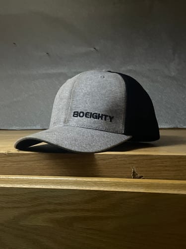 Customer's heather gray 80Eighty Melange Trucker Hat with a black embroidered logo, seen from the side on a wooden surface.