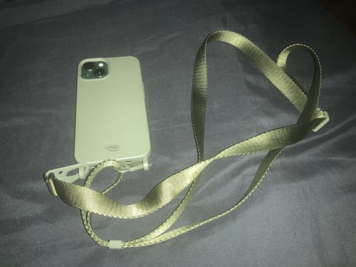 Cliente exibe a Alça Aurora verde musgo conectada a uma capa de celular, com a alça formando um coração.