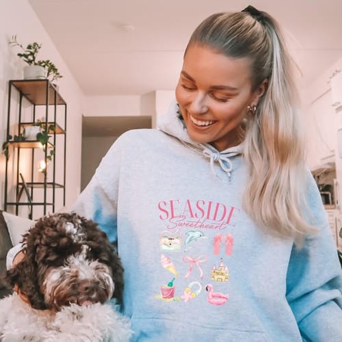 Reviewer smiling while wearing the light gray Seaside Sweetheart Hoodie next to a fluffy dog.