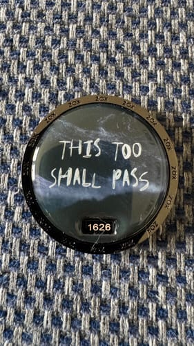 Customer's 'This Too Shall Pass' coin with handwritten-style text, resting on a blue patterned fabric.