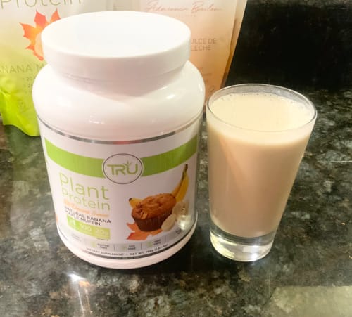 Customer's tub of Banana Maple Muffin flavored Protein Complete next to a prepared smoothie in a glass.