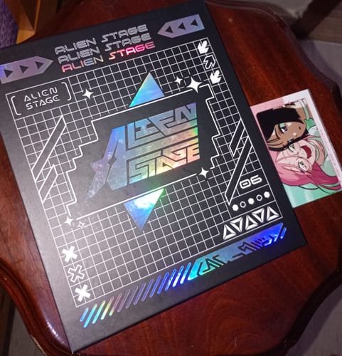 Customer's Alien Stage Card Collection Book with a holographic cover on a table, beside a character card freebie.
