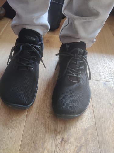 Ein Kunde trägt die schwarzen Wish Ⅰ - Barfußschuhe auf einem Holzboden.