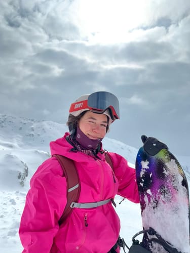 Reviewer in a bright rose Snow Anorak, holding a snowboard on a snowy, overcast day.