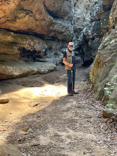 Reviewer standing with a walking stick on a dirt path surrounded by large, weathered rock formations.