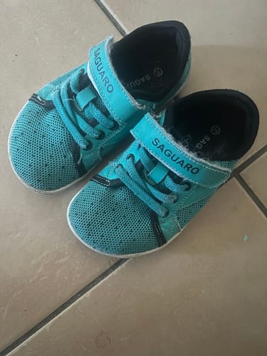 Ein Kunde zeigt ein Paar türkisfarbene Kinder Luck I Barfußschuhe mit Klettverschluss auf einem Fliesenboden.