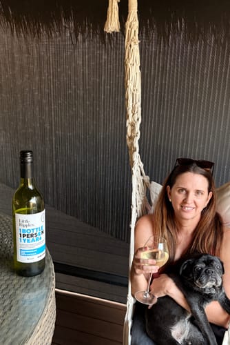 Customer on a patio holding a glass of white wine and a small dog, with a bottle of Sauvignon Blanc on the table.