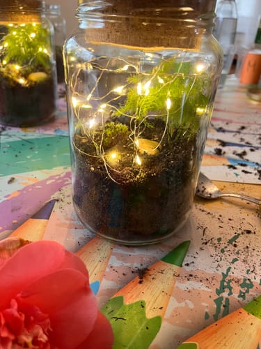 Kunde zeigt den fertigen Flaschengarten. Das Glas ist mit Erde, Pflanzen und einer leuchtenden Lichterkette gefüllt.