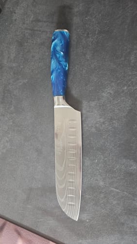 Ein Kunde zeigt das Shiburu Santoku-Messer, das auf einer dunklen Oberfläche liegt und einen blauen Harzgriff sowie eine gemusterte Klinge hat.
