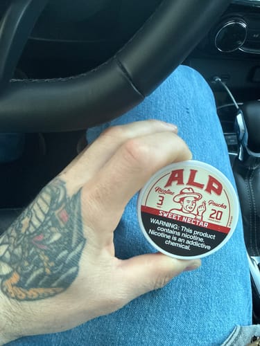 A reviewer's tattooed hand holding a round tin of Sweet Nectar Nicotine Pouches while sitting in a car.