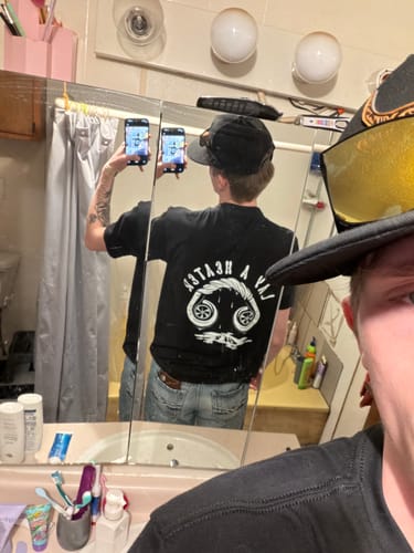 Customer taking a mirror selfie to show the back of the black HEATER TEE in a bathroom.
