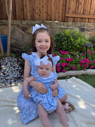 Reviewer wearing the blue floral dress, holding a baby in a matching smocked outfit outdoors.