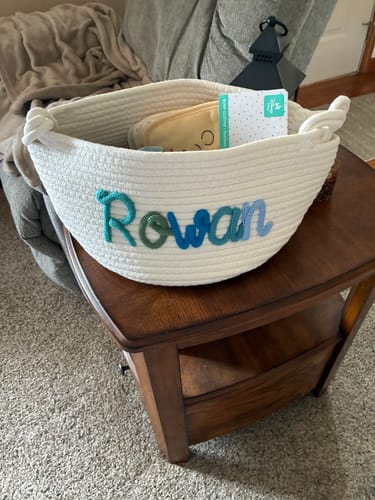 Customer's white personalized knitted basket with the name 'Rowan' in multicolor yarn, sitting on an end table.