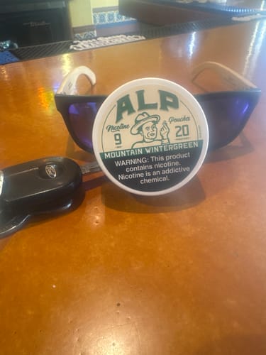 Customer's container of Mountain Wintergreen Nicotine Pouches on a wooden bar next to sunglasses and keys.