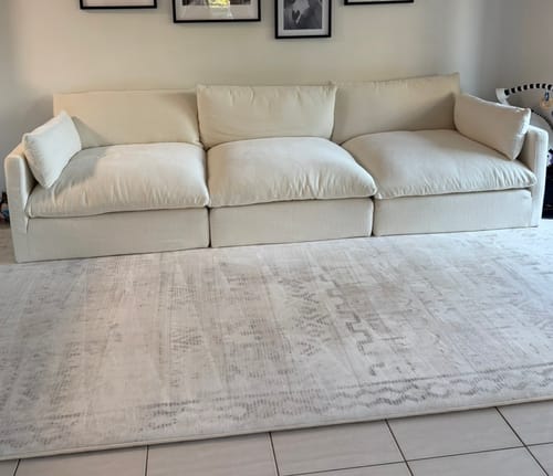 Customer's large Mellow Mat in Sandstone Haze laid out in a living room in front of a sofa.