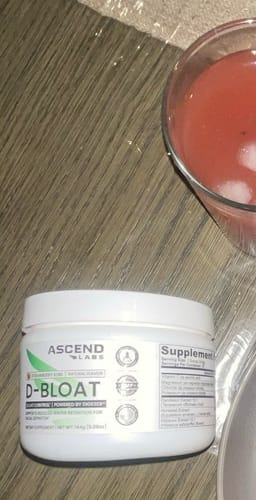 Customer's white tub of D-BLOAT powder next to a glass of the prepared reddish drink on a table.
