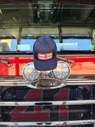 Customer's navy blue SNAPBACK HOSE HAT with an 'ENG 41 INE' patch resting on the grille of a red fire truck.