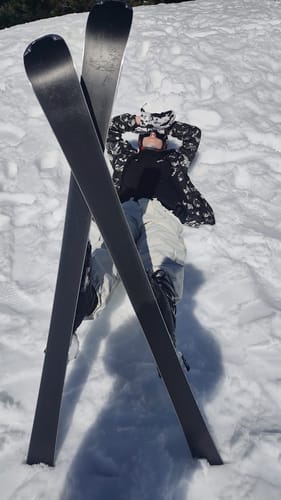 A reviewer lies in the snow wearing the beige baggy snow pants, with skis crossed in the foreground.