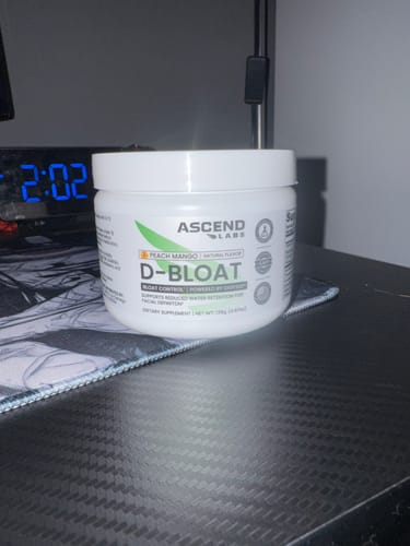 Reviewer's white tub of D-BLOAT powder sitting on a dark desk, next to a mousepad.