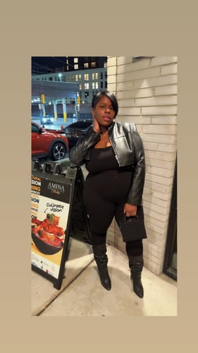 Reviewer models the black NW Square Neck Jumpsuit Curve, layered with a cropped jacket and boots on a city street at night.