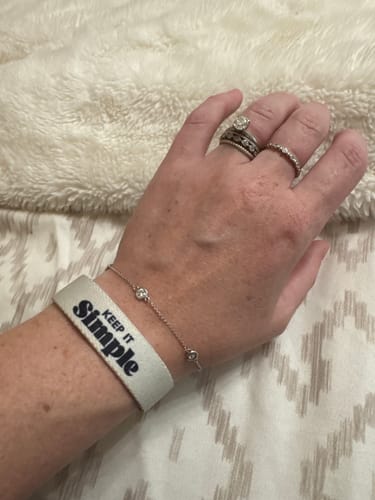Reviewer wearing the white fabric 'Keep it Simple' bracelet on their wrist, resting on a bedspread.