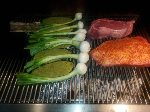 Dos Rib Eye Cowboy Steaks de un cliente en la parrilla, crudos y listos para cocinarse con cebollas y nopales.