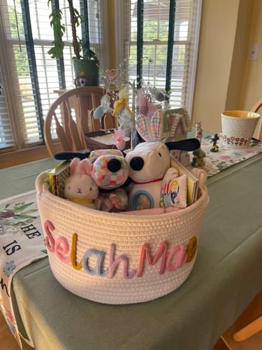 Customer's personalized white knitted basket with the name "Selah Mae" in colorful yarn, filled with toys on a table.