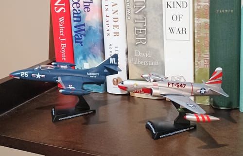 Reviewer's dark blue F9F Panther model on a stand, displayed on a bookshelf next to another model jet.