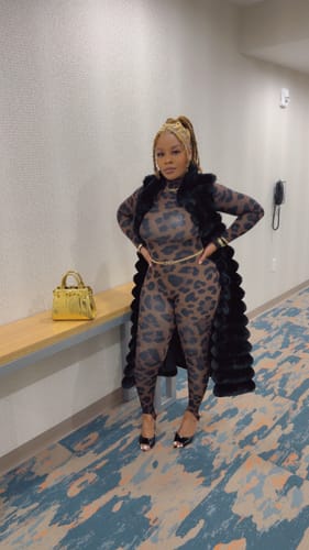 Reviewer standing in a hallway wearing the form-fitting Naked Print Stirrup Jumpsuit with a black vest.