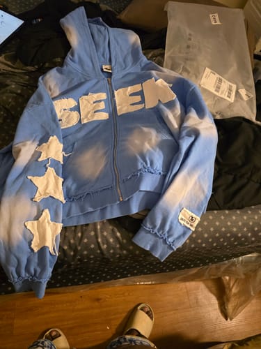 A customer's blue tie-dye VISIONZ SWEATS zip-up hoodie with white lettering and star patches on the sleeve, laid out on a bed.