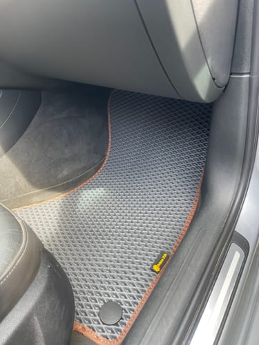 Recenzent pokazuje idealnie dopasowany szary dywanik samochodowy z brązowym obszyciem po stronie pasażera.