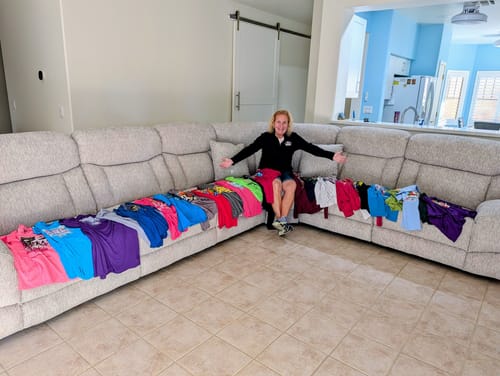 Customer sitting on a couch with many colorful t-shirts laid out, ready to be made into a Project Repat T-Shirt Quilt.