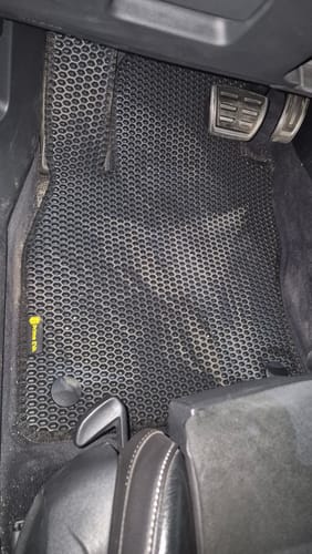 Recenzent pokazuje dopasowany, czarny dywanik samochodowy zainstalowany w aucie od strony kierowcy.