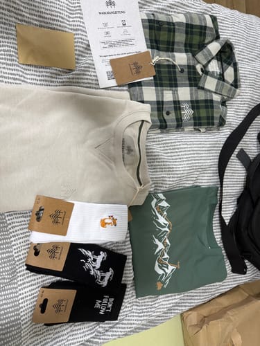Kunde zeigt den Inhalt der Holzbrüder Wundertüte: ein Sweatshirt, ein kariertes Hemd, ein T-Shirt und Socken auf einem Bett.