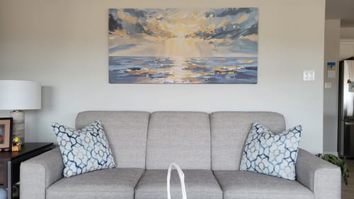 Reviewer's 'When Light Breaks Through' canvas hanging on a large wall above a grey sofa in a living room.