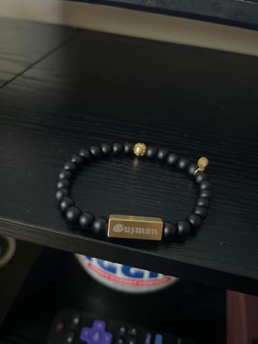 Customer's matte black bead bracelet with a gold rectangle bead engraved with 'Guzman' on a black shelf.