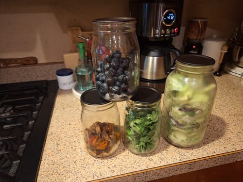 Customer's mason jars sealed with black Mason Genie lids, holding fresh produce and prepared food on a kitchen counter.