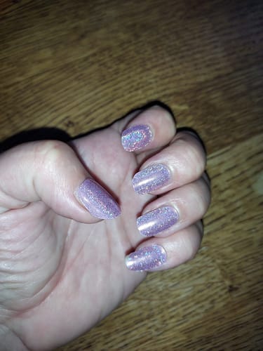 A reviewer's hand showing their long nails with sparkly Pink Glitz gel polish against a wood surface.
