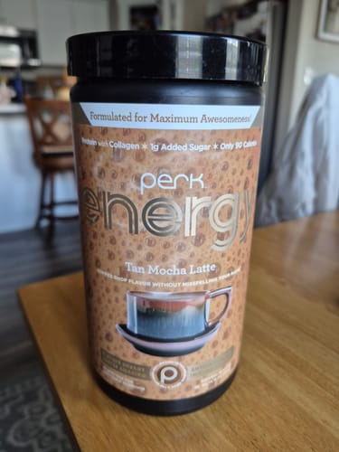 Customer's black and brown canister of Perk Energy drink mix sitting on a wooden table in a kitchen.